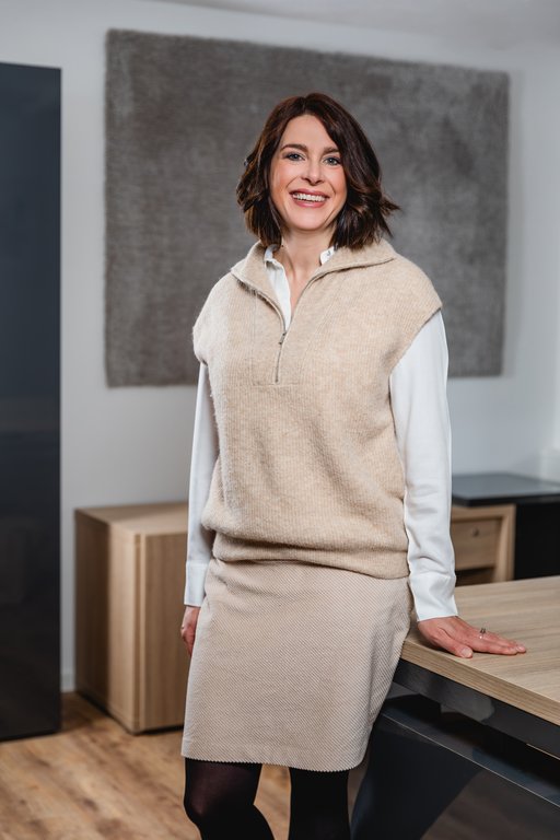 Frau lächelt, steht und lehnt an einem Tisch. Sie trägt einen cremefarbenen Pullover über einem weißen Hemd. Umgebung ist ein heller Raum mit Holzboden und Büroeinrichtung.