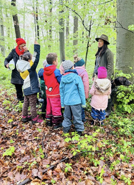 Eine Gruppe von Kindern steht im Kreis um eine Frau im Wald. Einige Kinder tragen bunte Jacken und Mützen, während die Frau etwas erklärt. Feuchter Waldboden umgibt sie.