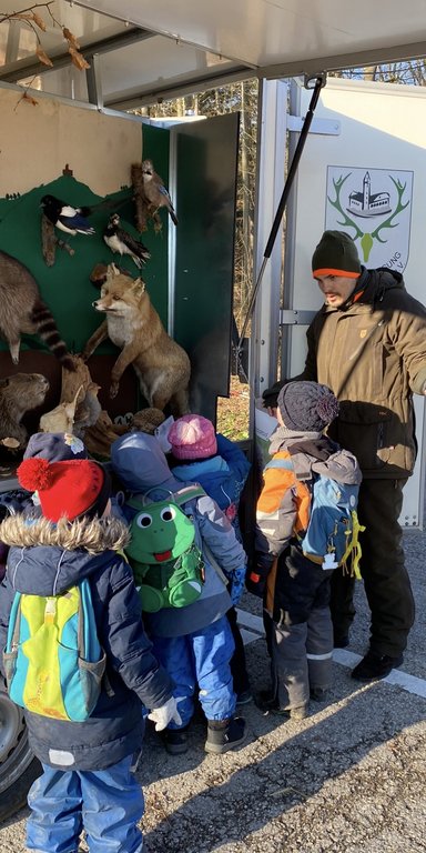 Ein Mann erklärt einer Gruppe Kinder ausgestopfte Tiere in einem Anhänger. Die Kinder tragen bunte Winterkleidung. Im Hintergrund ist ein Logo mit einem Geweih auf dem Anhänger.