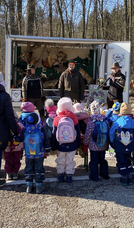 Kindergruppe steht vor einem offenen Anhänger mit Tierpräparaten, während drei Erwachsene, in Outdoor-Kleidung, erklären. Der Hintergrund zeigt einen winterlichen Wald. Text: "Jagd erleben".