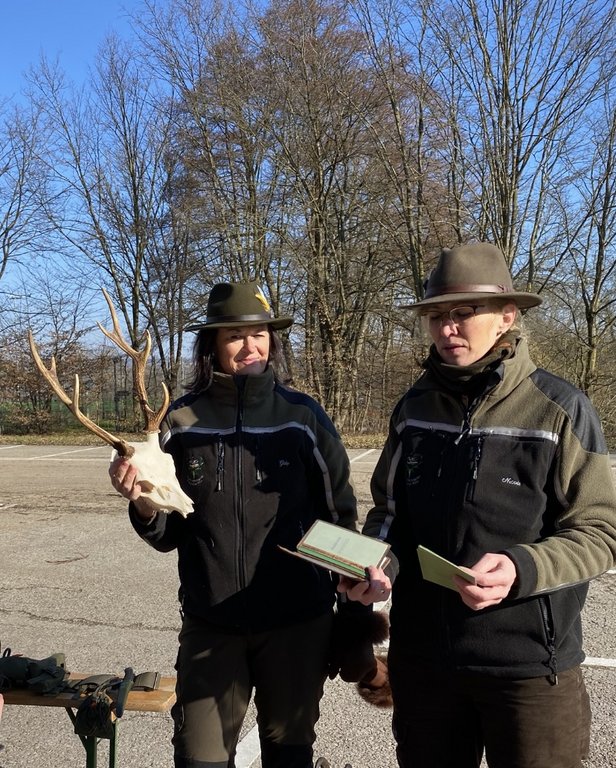 Zwei Personen in Forstkleidung halten einen Hirschschädel und Notizblöcke in den Händen, stehend auf einem Parkplatz, umgeben von Bäumen an einem sonnigen Tag.