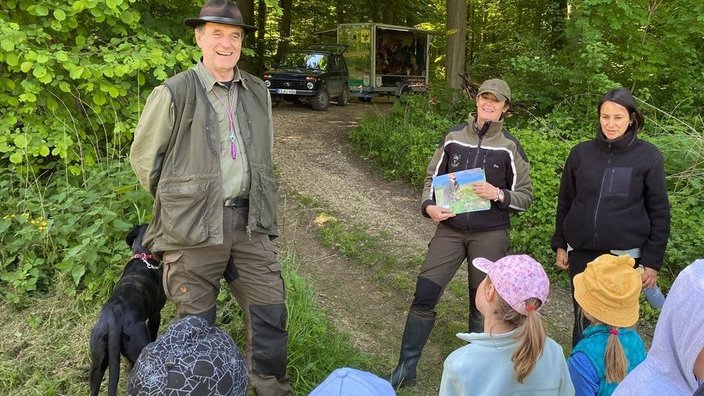 Ein Mann mit Hund steht lächelnd neben einer Frau, die ein Buch hält, umgeben von Kindern in bunten Mützen, im grünen Wald; im Hintergrund ein Fahrzeug.