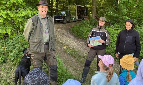 Ein Mann mit Hund steht lächelnd neben einer Frau, die ein Buch hält, umgeben von Kindern in bunten Mützen, im grünen Wald; im Hintergrund ein Fahrzeug.