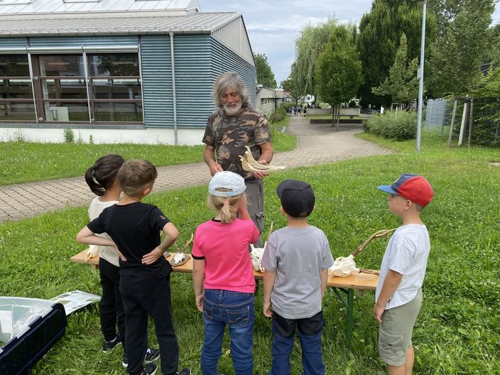 Ein Mann zeigt Kindern Knochen und Hörner auf einem Tisch im Freien. Sie befinden sich vor einem Gebäude mit Rasen und Bäumen umgeben.
