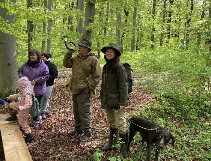 Ein älterer Mann bläst ein Jagdhorn, während er mit mehreren Menschen, darunter ein Kind und ein Hund, in einem grünen Wald steht. Alle sind warm gekleidet und lächeln.
