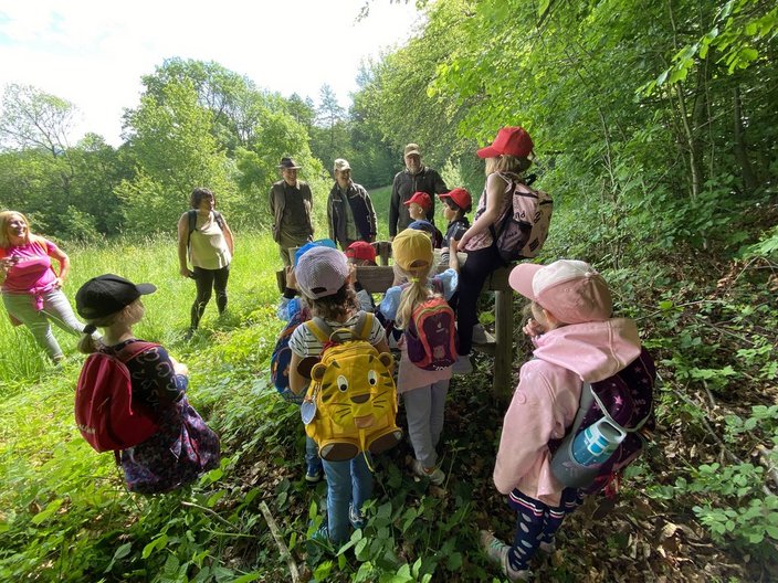 Eine Gruppe von Kindern mit bunten Rucksäcken steht im Wald und hört aufmerksam mehreren Erwachsenen zu, die Informationen geben. Die Umgebung ist grün und bewaldet.
