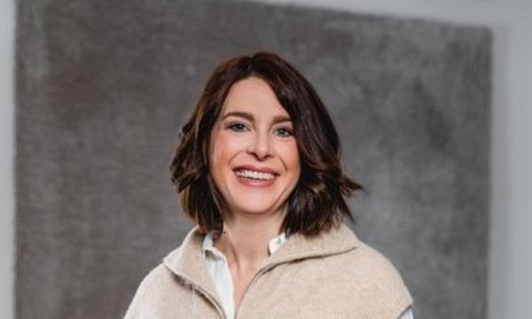 Frau lächelt, steht und lehnt an einem Tisch. Sie trägt einen cremefarbenen Pullover über einem weißen Hemd. Umgebung ist ein heller Raum mit Holzboden und Büroeinrichtung.