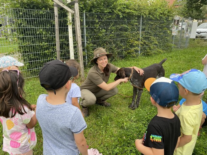 Hund mit braunem Fell steht ruhig, von Frau in grüner Uniform gestreichelt, umgeben von aufmerksam zuschauenden Kindern; im Hintergrund grüner Garten mit Zaun.
