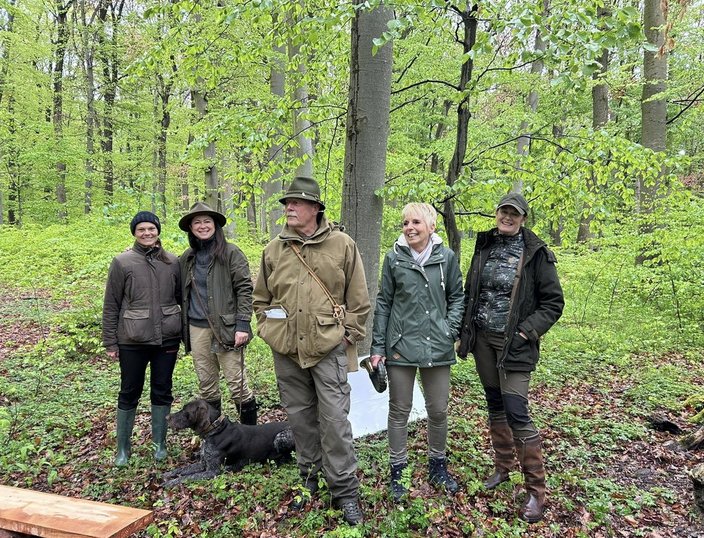 Fünf Personen stehen lächelnd in einem grün belaubten Wald, eine davon hält die Leine eines Hundes. Sie tragen Outdoor-Kleidung, während sie zwischen den Bäumen posieren.