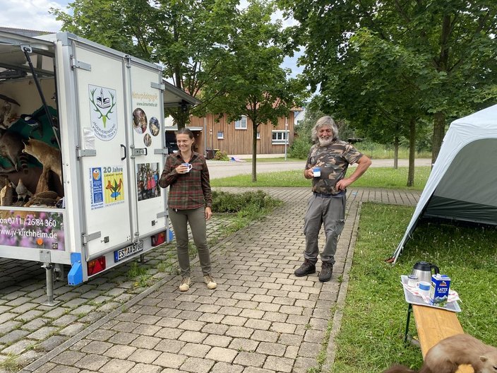 Ein Mann und eine Frau trinken Kaffee neben einem Anhänger mit Tiermodellen. Der Hintergrund zeigt Bäume, eine Wiese und ein Zelt. Texthinweise: "Naturpädagogik," "20 Jahre," "www.jv-kirchheim.de".