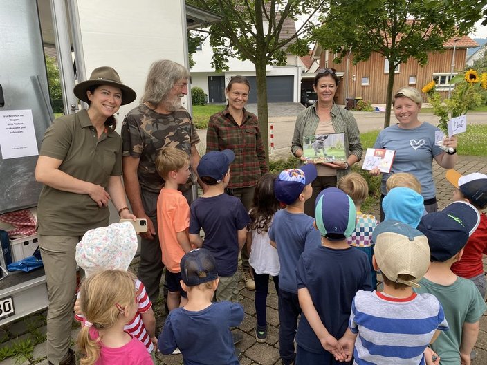 Eine Gruppe Erwachsener hält Bücher vor und lächelt, umgeben von Kindern mit Kopfbedeckungen, im Freien auf einem gepflasterten Gehweg. Im Hintergrund sind Häuser und Bäume sichtbar.

Text: "Vor Betreten des Wagens: Bitte Schuhe und Kleidungen untersuchen. Danke."