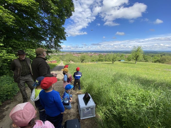 Kinder mit bunten Mützen stehen auf einem Waldweg. Sie schauen in die Ferne, wo ein Hubschrauber fliegt. Zwei Erwachsene beobachten sie. Eine Wiese und Bäume umgeben die Szene.
