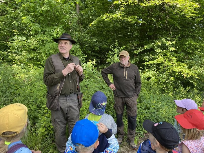Ein Mann spricht zu einer Gruppe von Kindern. Sie tragen bunte Hüte. Im Hintergrund sind dichtes grünes Laub und Bäume. Der Mann trägt einen Hut und eine Weste.