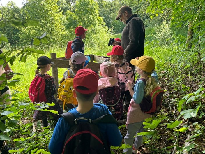 Eine Gruppe von Kindern mit Rucksäcken steht in einem Wald und hört einem Erwachsenen mit Fernglas zu, der etwas erklärt. Umgeben von dichtem grünen Laub und einem sonnigen Feld.
