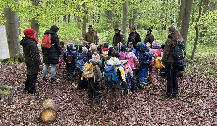 Kindergruppe mit bunten Rucksäcken steht im Kreis, umgeben von Erwachsenen, die im Wald Informationen geben. Die Szene spielt sich in einem grünen, dicht bewaldeten Bereich ab.