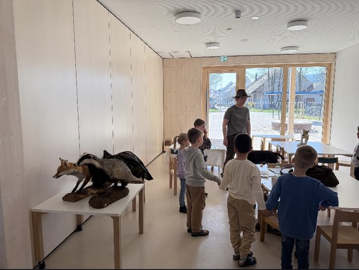 Ein ausgestopfter Fuchs und eine Gans stehen auf einem Tisch. Kinder umringen eine Person in einem hellen Raum mit Holzwänden, während sie einem Vortrag lauschen. Draußen ist ein Spielplatz sichtbar.